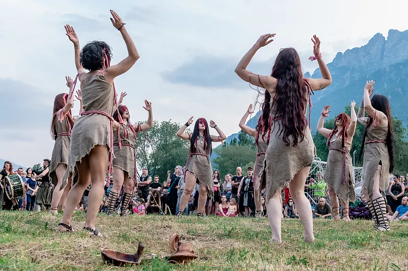 Gruppo di danzatori disposti in cerchio in un'azione coreografica all'aperto, con movimenti delle braccia sincronizzati. Indossano costumi naturali ispirati a tessuti grezzi, immersi in un paesaggio montano con pubblico sullo sfondo. Scena del laboratorio coreografico Fairy Circle dedicato alla danza rituale e alla connessione con il movimento collettivo