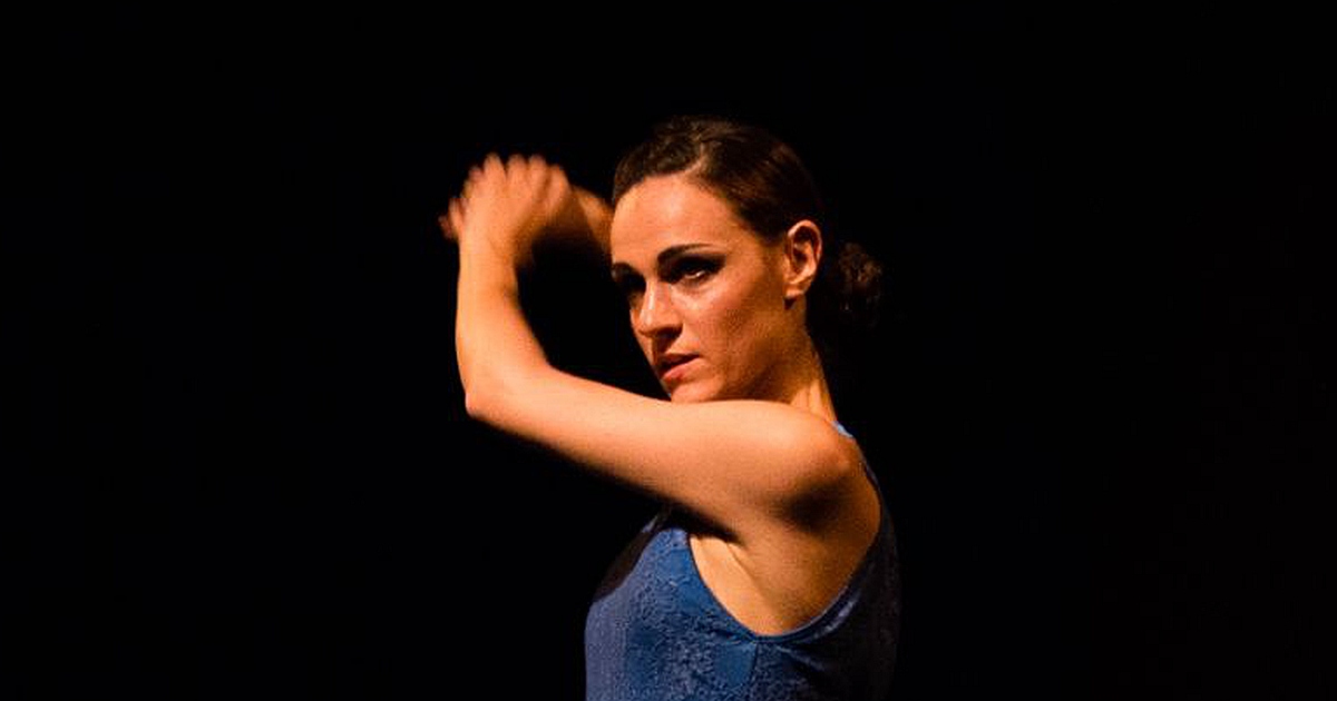 Ballerina di danza moderna in posa espressiva su sfondo scuro. Indossa un top blu senza maniche e una gonna fluida coordinata, con le braccia sollevate e lo sguardo intenso. L'illuminazione drammatica mette in risalto il movimento e la forza emotiva della performance