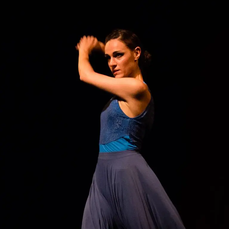 Ballerina di danza moderna in posa espressiva su sfondo scuro. Indossa un top blu senza maniche e una gonna fluida coordinata, con le braccia sollevate e lo sguardo intenso. L'illuminazione drammatica mette in risalto il movimento e la forza emotiva della performance