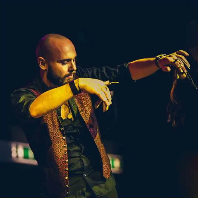 Gianluca Insalata, un uomo calvo e barbuto, con camicia nera e gilet tradizionale, danza energicamente sul palco con le braccia tese. Insegnante di Pizzica e Tammurriate