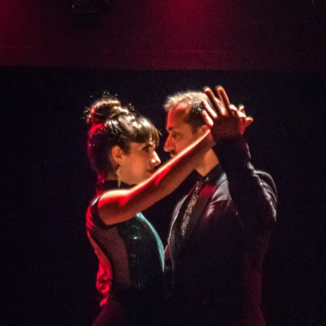 Lucila Prego e Roberto Angelica in un'intensa posa di Tango Argentino sotto luci rosse di scena, con uno sguardo ravvicinato e l'abbraccio alto tipico del tango.