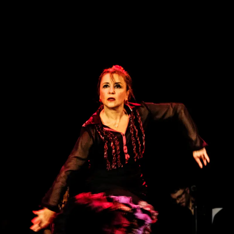 Sabina Todaro esegue una potente performance di flamenco, indossando un abito tradizionale nero con accenti rossi e viola. Le sue braccia sono sollevate in una posa espressiva e il suo volto intenso trasmette tutta la passione e la drammaticità della danza. Lo sfondo scuro mette in risalto la sua figura e l'energia del momento
